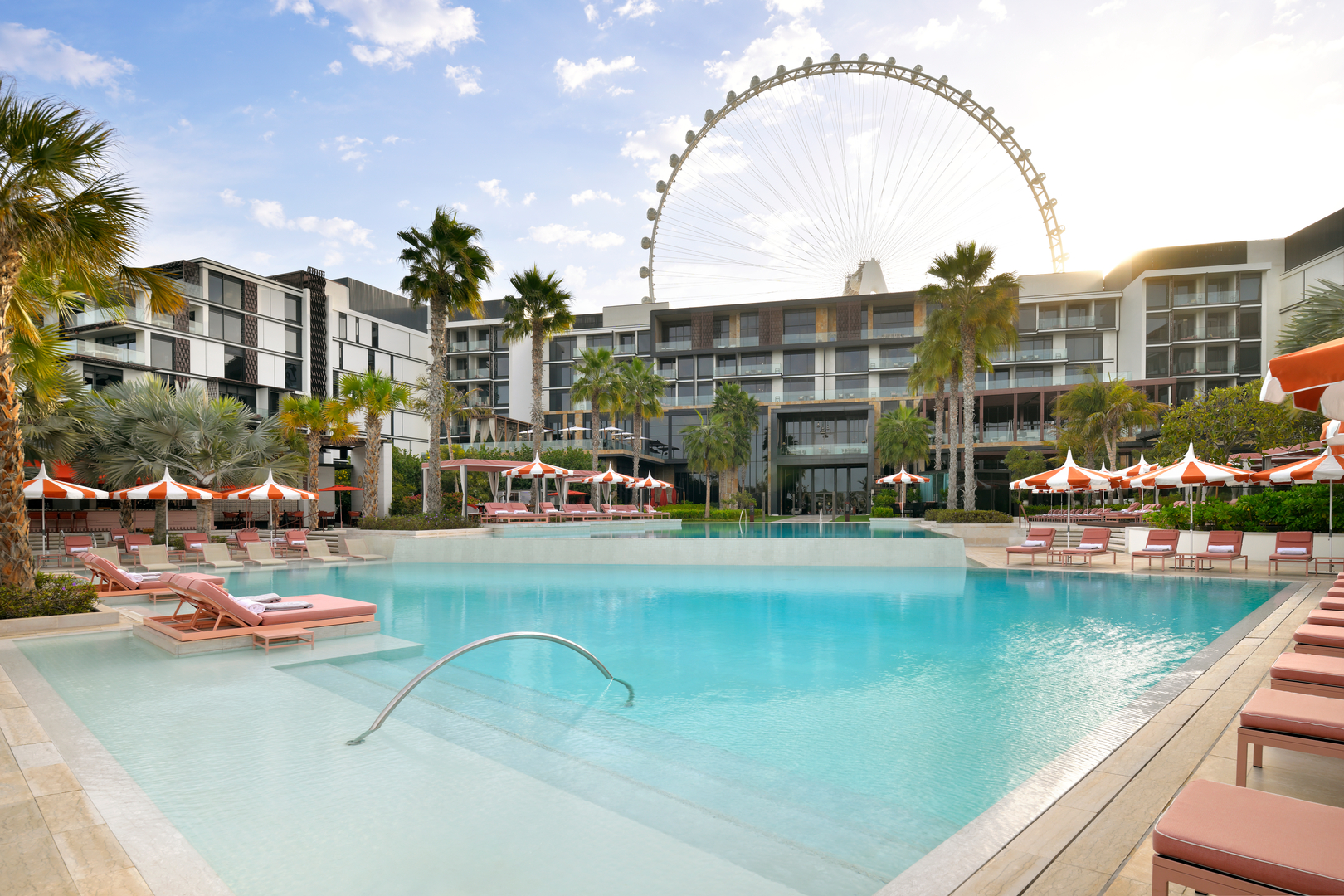 Sunny daytime view of a luxurious resort pool surrounded by palm trees, red-and-white umbrellas, and pink sun loungers. The Ain Dubai observation wheel stands tall in the background, with the modern resort building creating a stunning backdrop for this vibrant and relaxing setting.